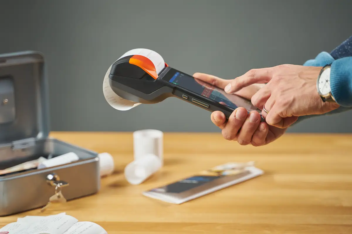 Hands feeding paper through a portable receipt printer on a wooden table.